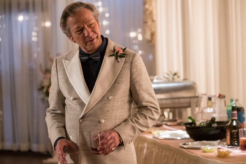 An older man with wry smile stands in formal beige suit with black shirt and bowtie and holds two glasses in wedding venue.