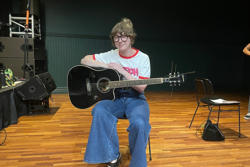 A woman with glasses, wearing jeans and a t-shirt sits with a black guitar in her lap. 