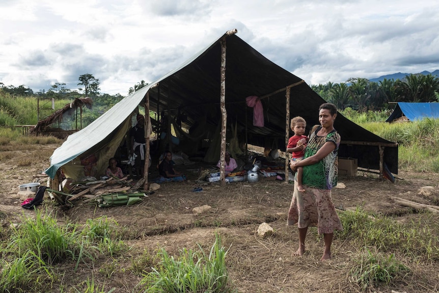 Wide shot of a woman standing holding her baby in front of a large tent.