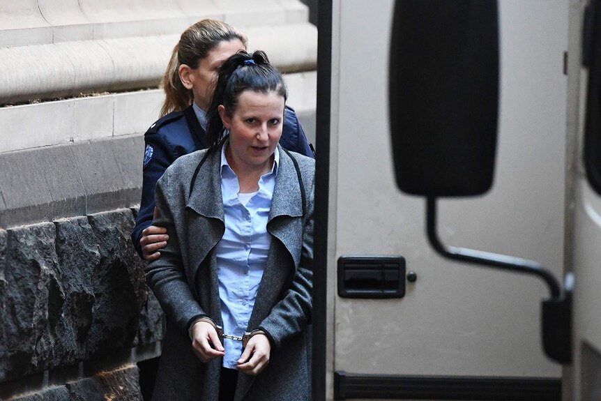 A young woman in a blue shirt and grey coat is escorted into a prison van.