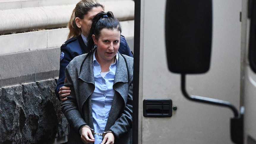 A young woman in a blue shirt and grey coat is escorted into a prison van.