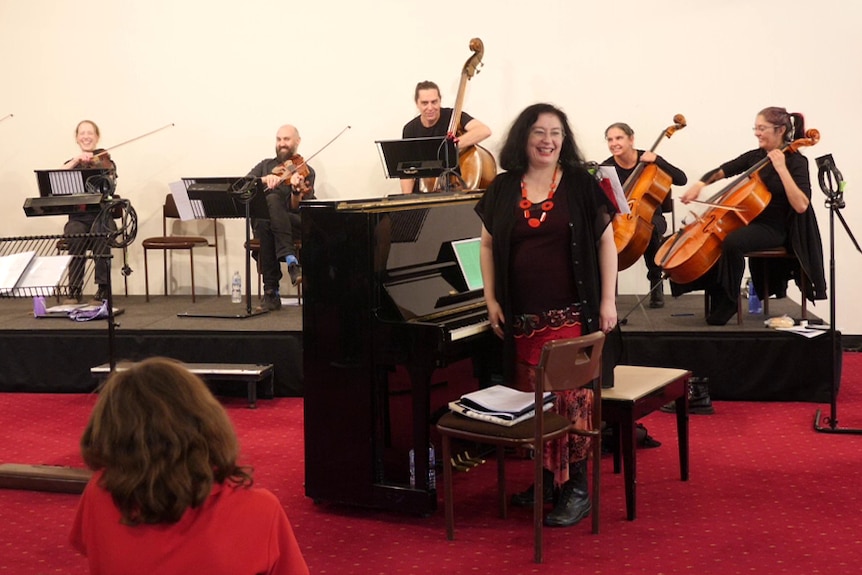 Musicican wearing black with woman in black and red in foreground in front of black upright piano.