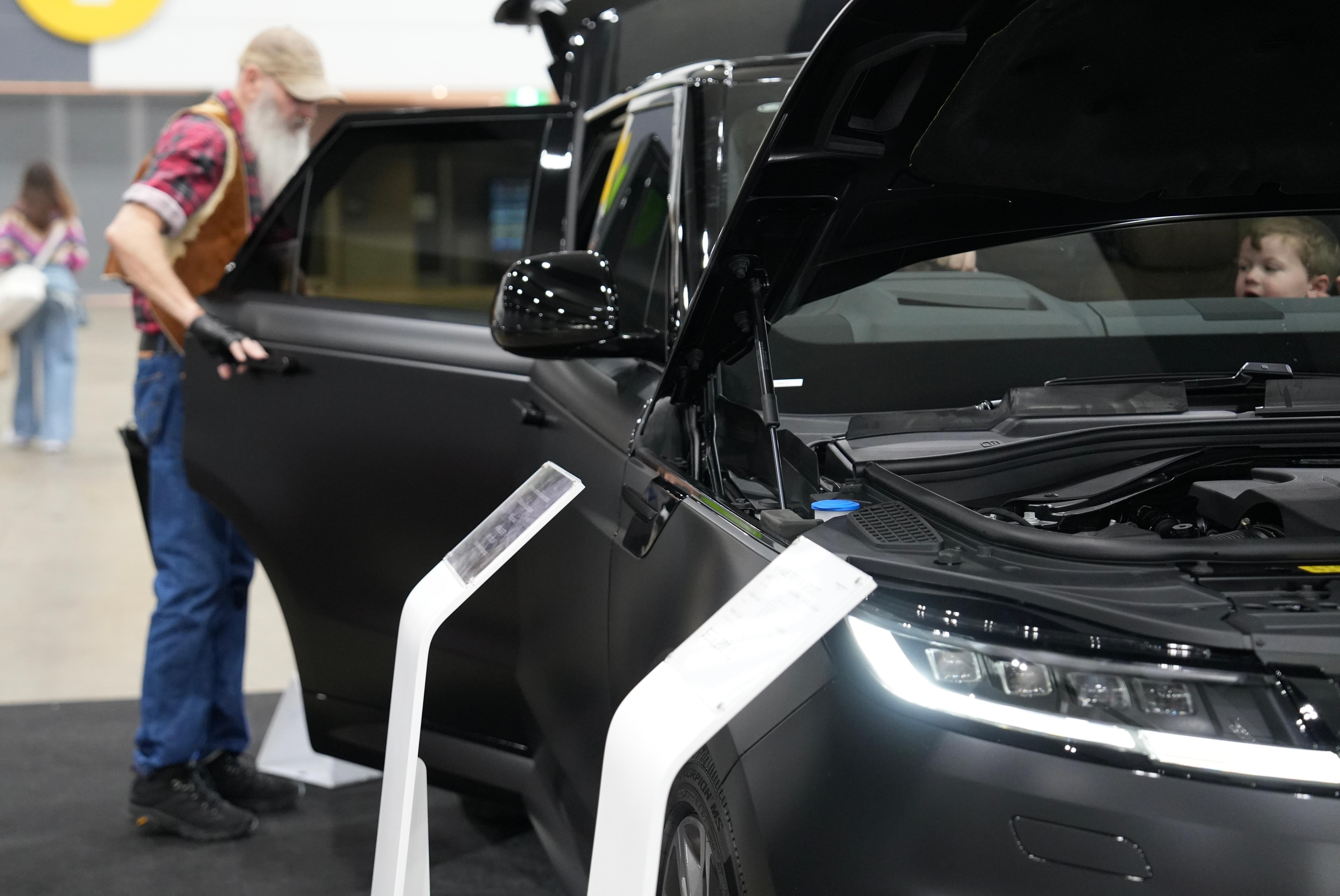 An older man with a long, white beard looks at a black car in a showroom.