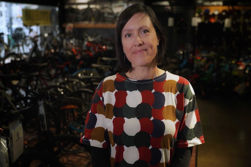 A dark haired woman in a multi-coloured top stands in front of bicycles in a shop.
