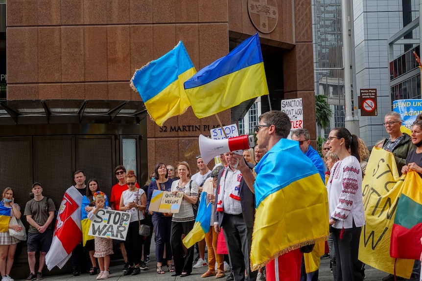 A man draped in a Ukrainian flag speaks through a megaphone, with several flags visible among the crowd behind him.