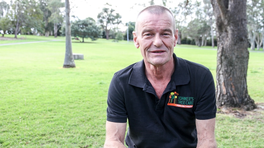 A man with short hair, wearing a dark shirt, sits in a park, smiling.