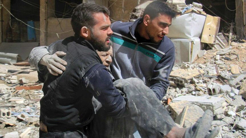 Two men carry and injured man in their arms past rubble.