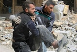 Two men carry and injured man in their arms past rubble.