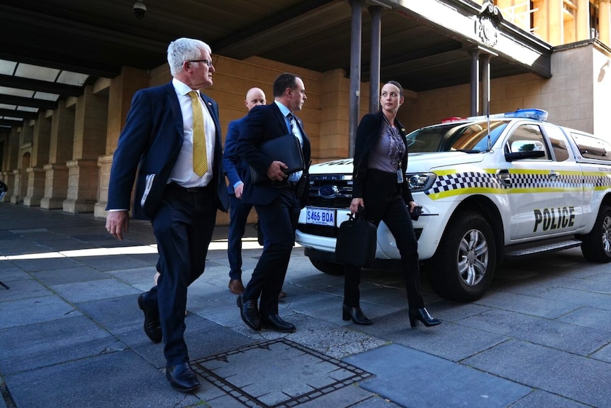 Three men and one woman wearing suits walking beside a police car. 
