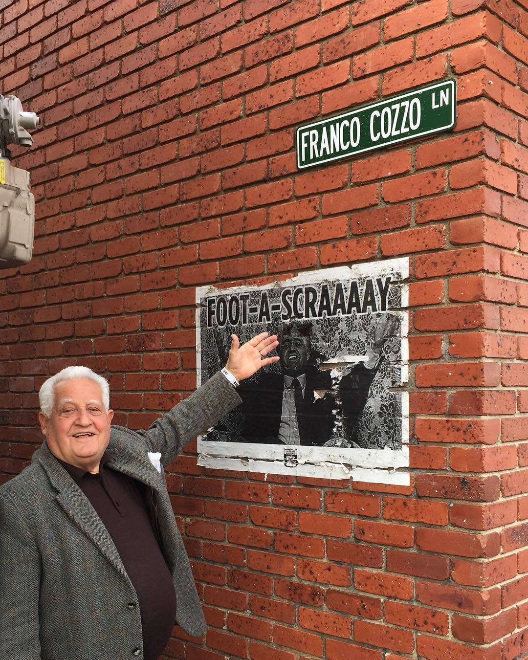 Franco Cozzo pointing to a poster with his face on it, beneath an unofficial lane signage reading Franco Cozzo lane.