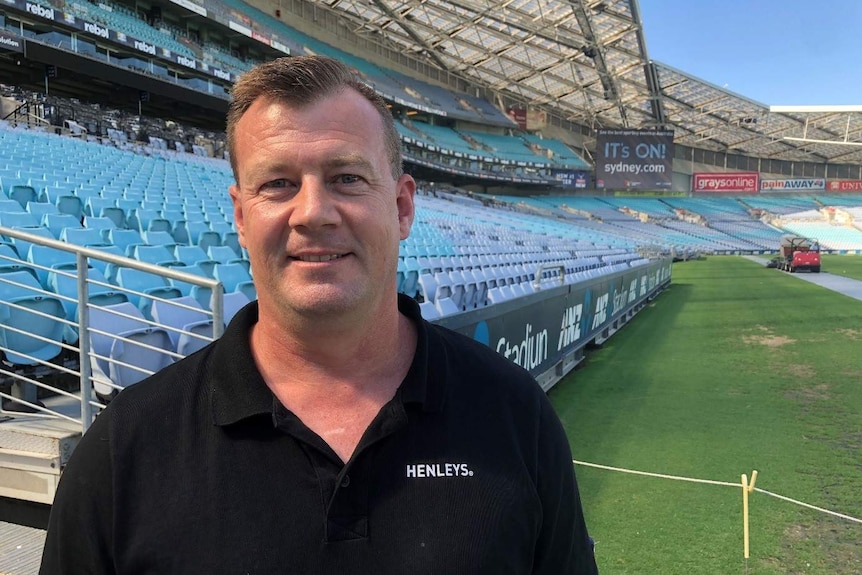 Dave Hendricks smiles at the camera inside the Olympic stadium, wearing a black polo shirt