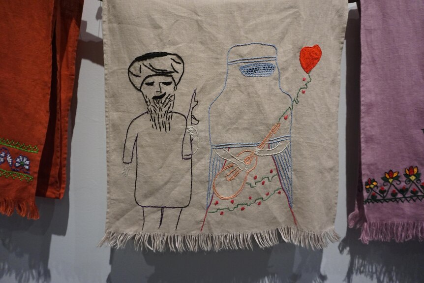 An embroidered scarf with an image of a woman in a burqa playing a guitar while a Taliban member looks at her.