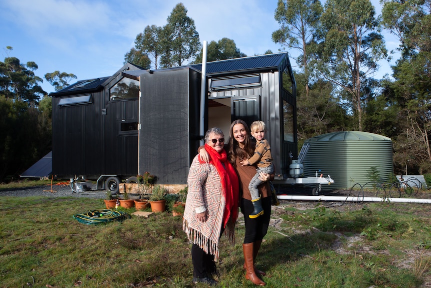 Two women and a toddler pose outside a mobile tiny house on a bush block.