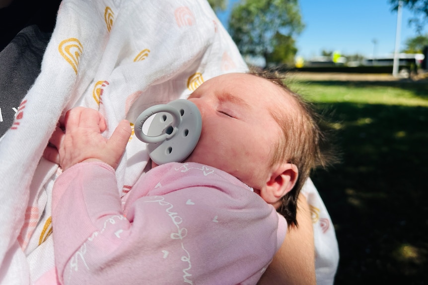 A six-week-old baby sleeping in her mums arms with a dummy in her mouth