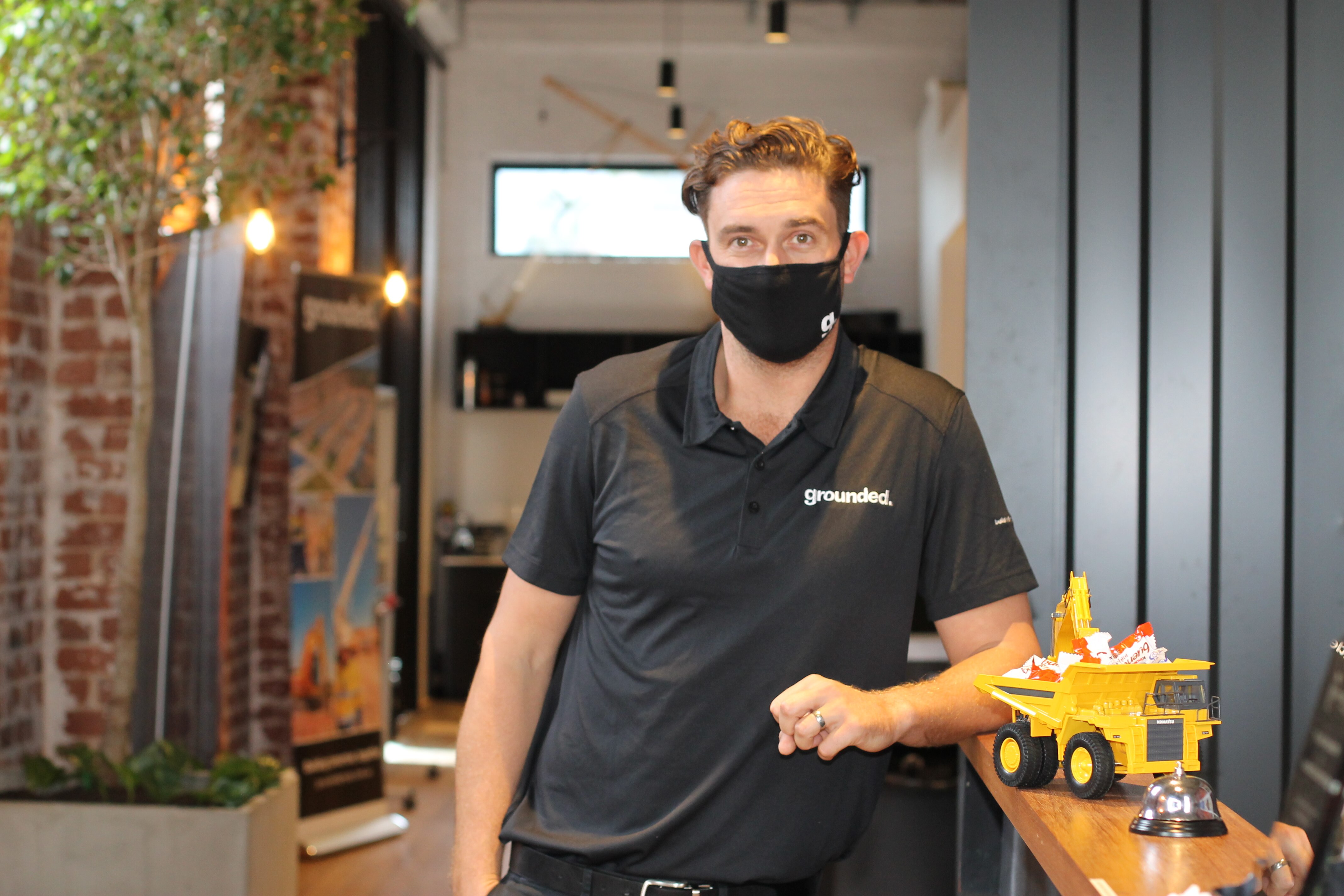 Courtney wears a black t-shirt with a white logo, also wearing a black face mask, standing inside an office.