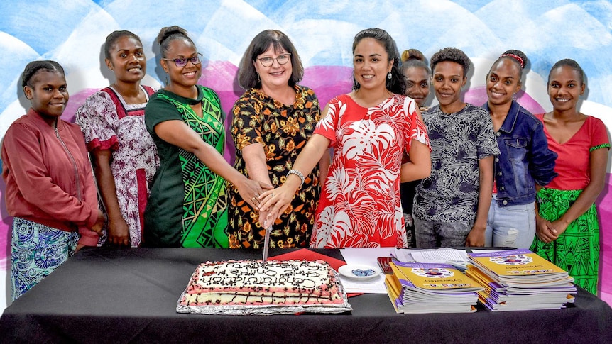 Nine women gather to perform a celebratory cake cutting. Three women in the centre each have their hand on the knife.