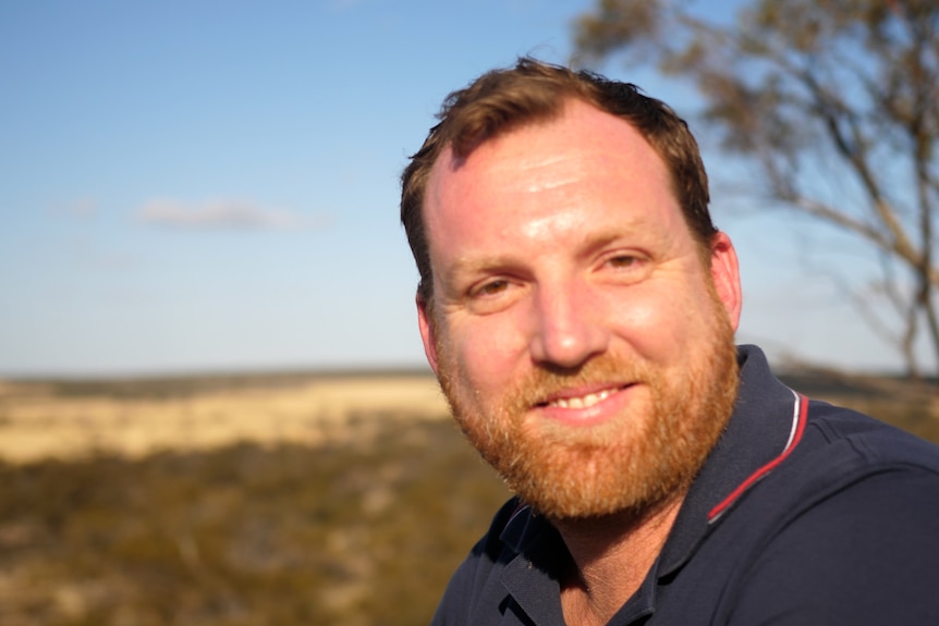 A man with red hair and a red beard smiles at the camera in front of a rural scene.