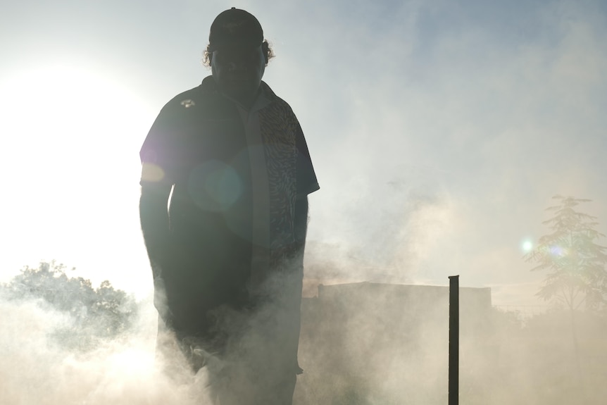 A man stands amongst smoking branches against the backdrop of the sun