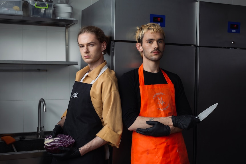 Two man in aprons stand in a kitchen looking at the camera. 