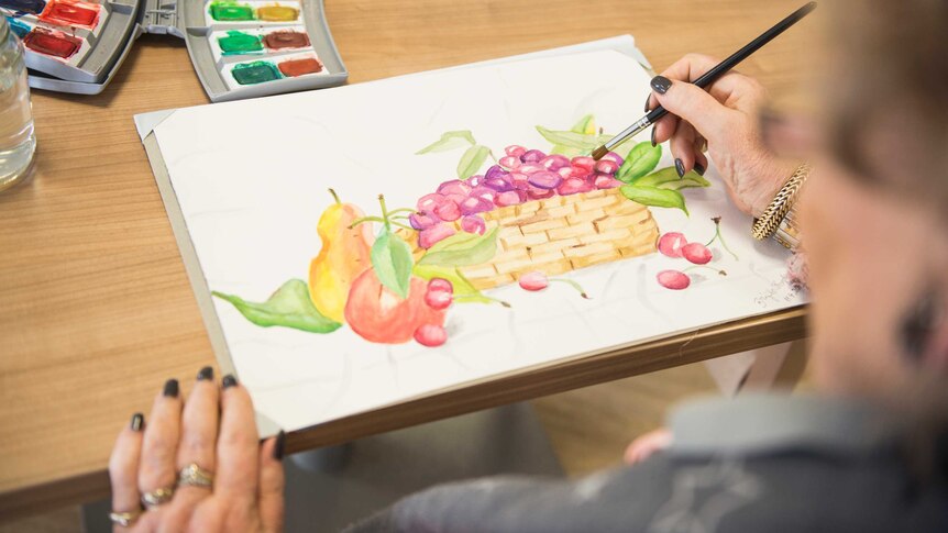 An older woman uses water colours to paint a picture of a fruit basket.