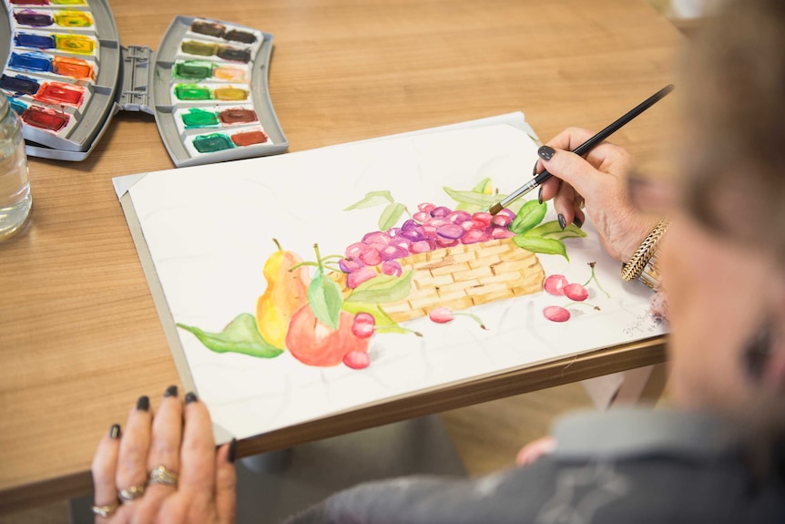 An older woman uses water colours to paint a picture of a fruit basket.