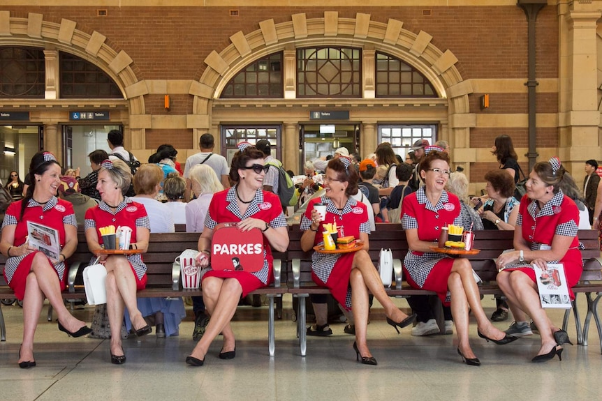 Women dressed in 1950s-styled diner waitress uniforms sitting on train station benches.
