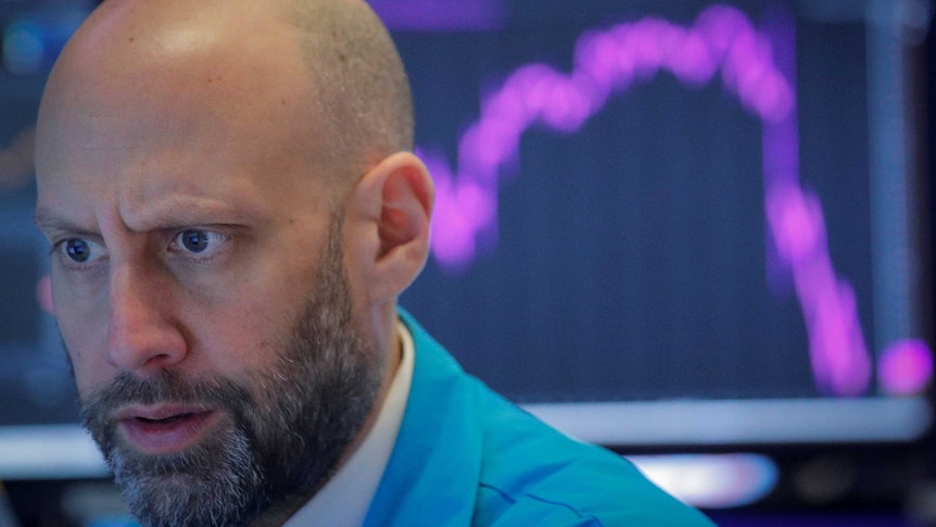 A man sits in front of a monitor with a graph showing a sharp drop.