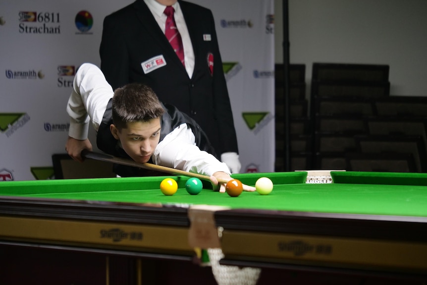 A young boy wearing a white shirt and black vest leans over to pot a snooker shot on a green table