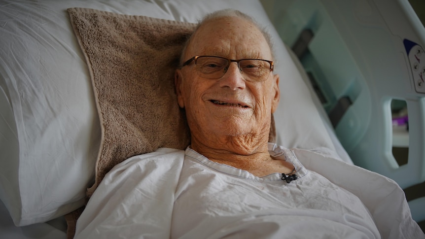 A man lies in a hospital bed and smiles at the camera