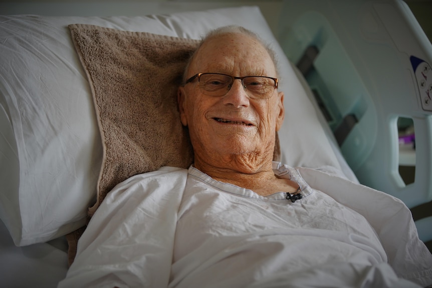 A man lies in a hospital bed and smiles at the camera