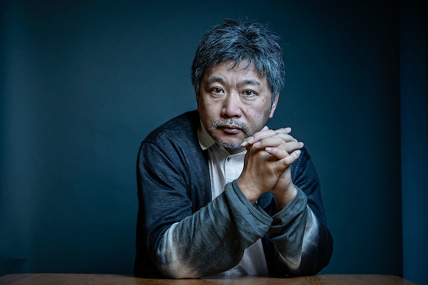 A man with short greying hair sits in front of blue grey backdrop and rests on table with hands clasped together.