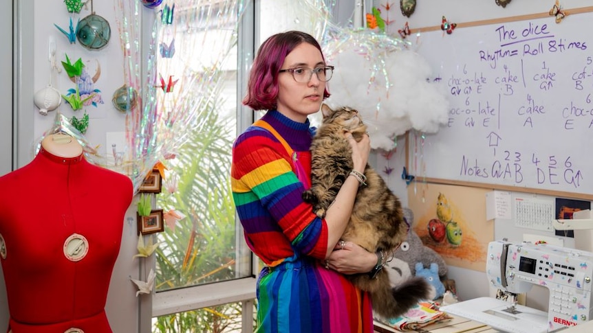 A young woman with short pink hair, glasses and rainbow jumper cuddles a cat in a sewing room.
