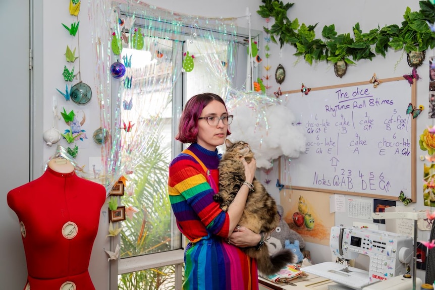 A young woman with short pink hair, glasses and rainbow jumper cuddles a cat in a sewing room.