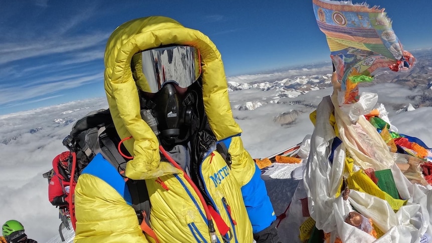 Colac's Luke Rollnik stands on the summit of Mount Everest wearing high-altitude face coverings and holding a flag. 