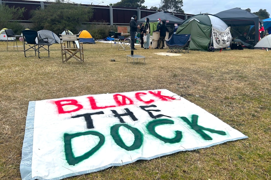 A Block the Dock banner written in paint lies on the ground. Tents in the background.