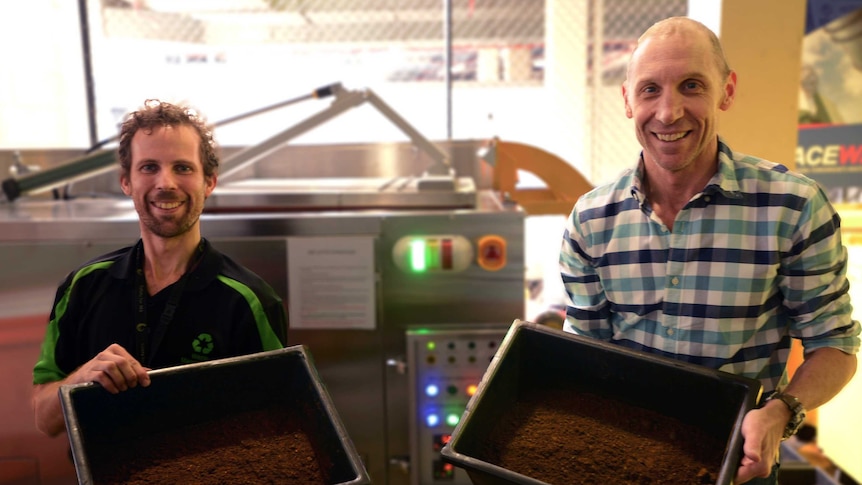 Two men holding containers of soil in front of a dehydrator.