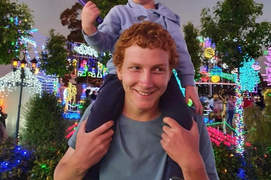 A young man with a child on his shoulders looks directly into the camera.