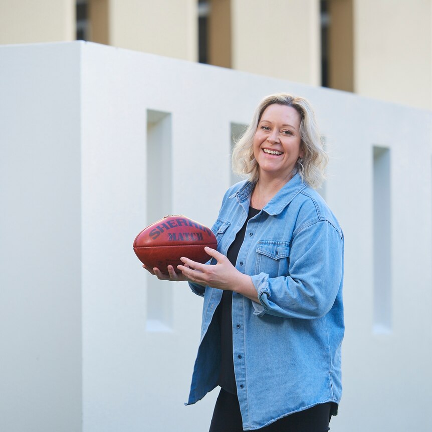 Andrea Gibbs, outdoors, holds a Sherrin AFL football and smiles broadly at the camera.