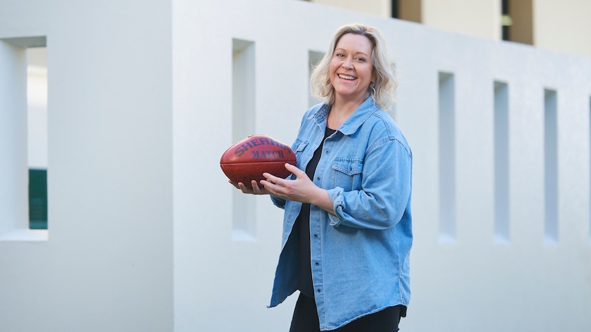 Andrea Gibbs, outdoors, holds a Sherrin AFL football and smiles broadly at the camera.