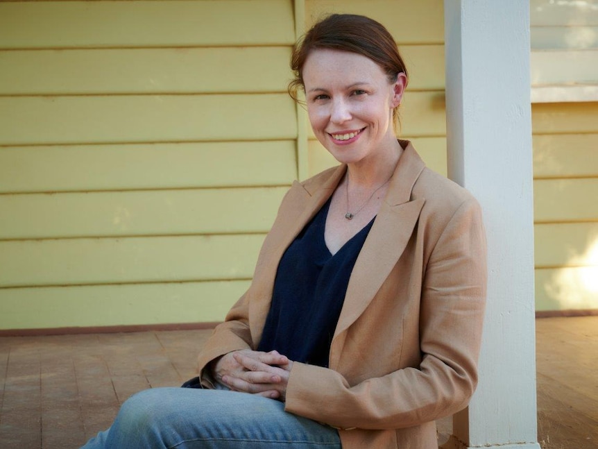 Ali Cupper sitting on a verandah, leaning on a white pole in a beige blazer.