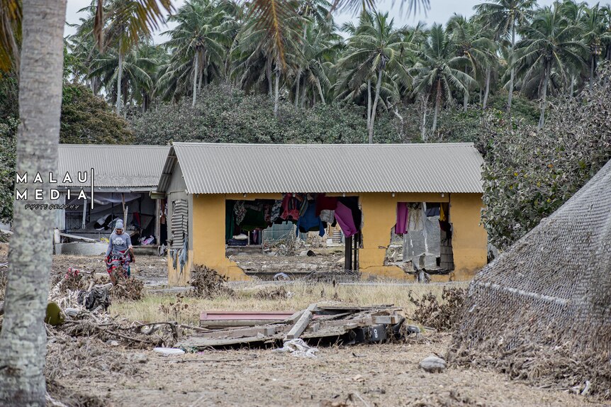 Outdoor photograph of home structures left in ruin after tsunami in Tonga. Woman stands by dilapidated house