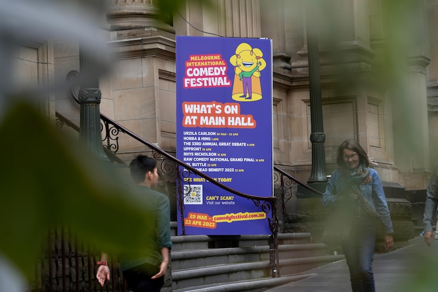 A sign for the Melbourne International Comedy Festival is seen through trees which are in soft focus.