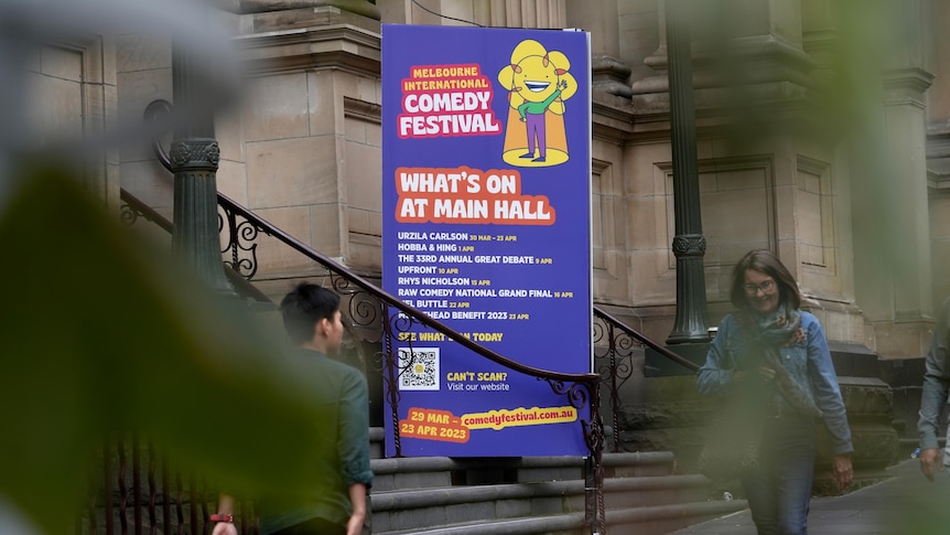 A sign for the Melbourne International Comedy Festival is seen through trees which are in soft focus.