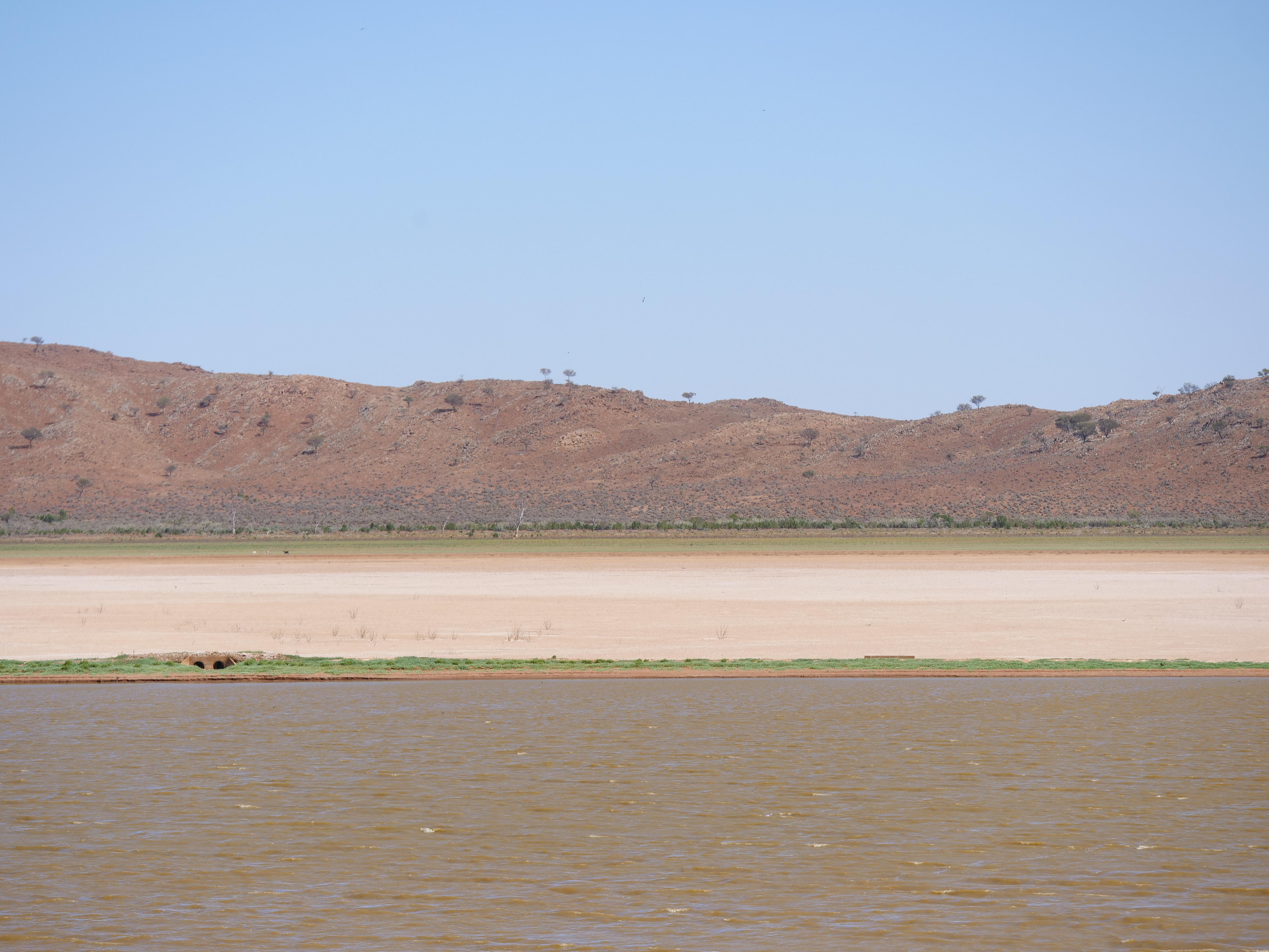 Uma extensão de água é mostrada em primeiro plano, com um trecho de leito de lago seco e montanhas sob um céu claro ao fundo.