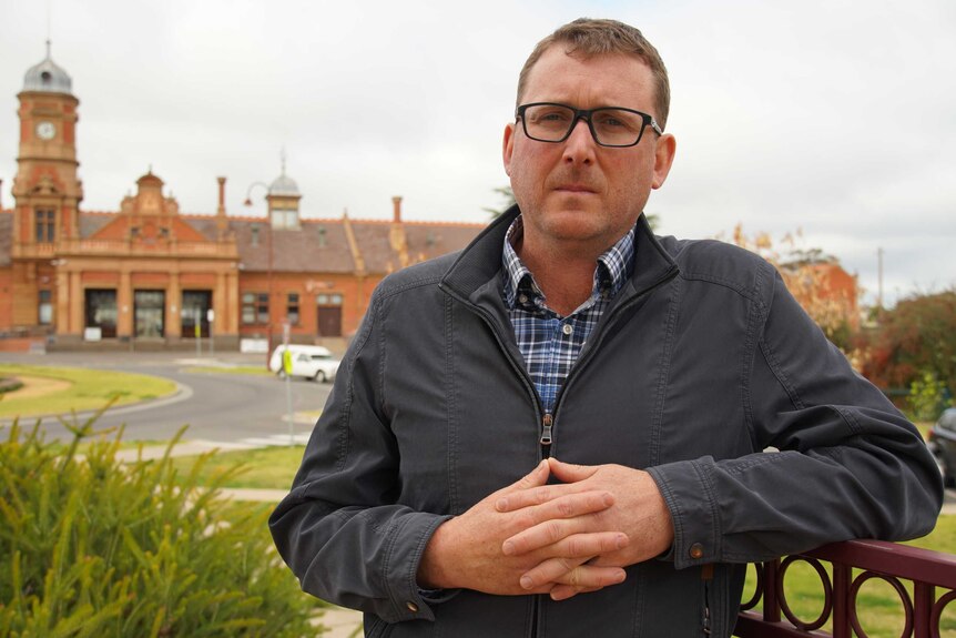 A man in his late 30s stands with his hands clasped with an od red brick building in the background.