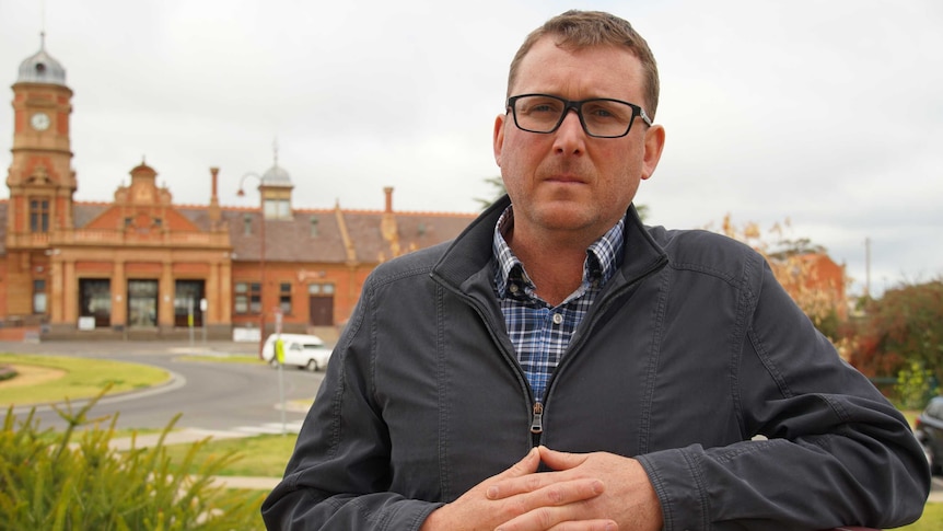 A man in his late 30s stands with his hands clasped. An old red brick building is in the background.