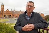 A man in his late 30s stands with his hands clasped. An old red brick building is in the background.