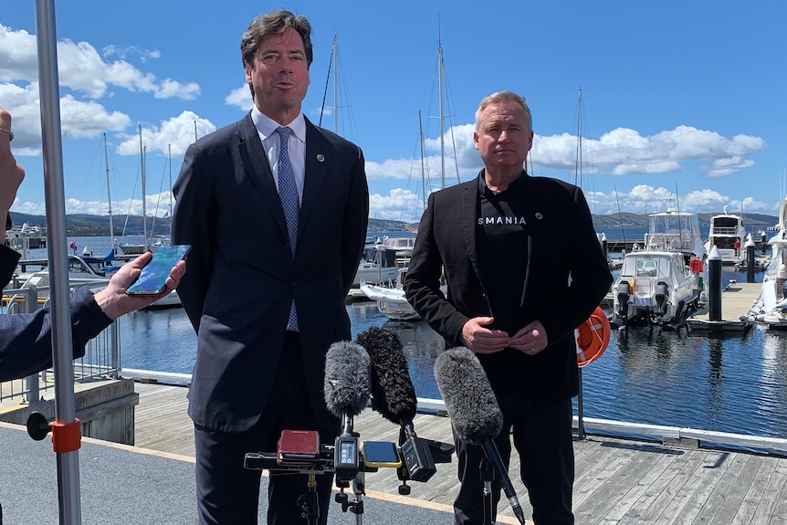 Two men in front of microphones on Hobart's waterfront in front of docked boats.