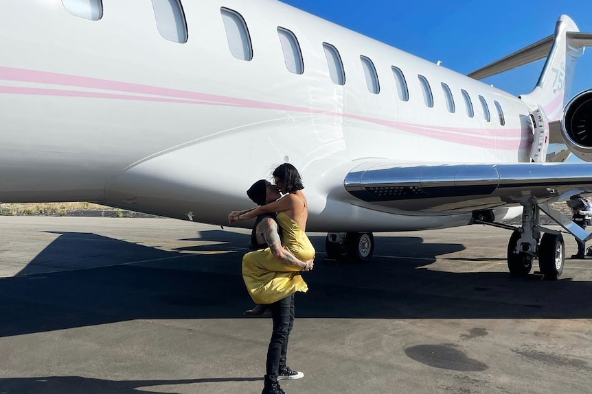 Travis lifts Kourntney as they kiss on the tarmac in front of a plane