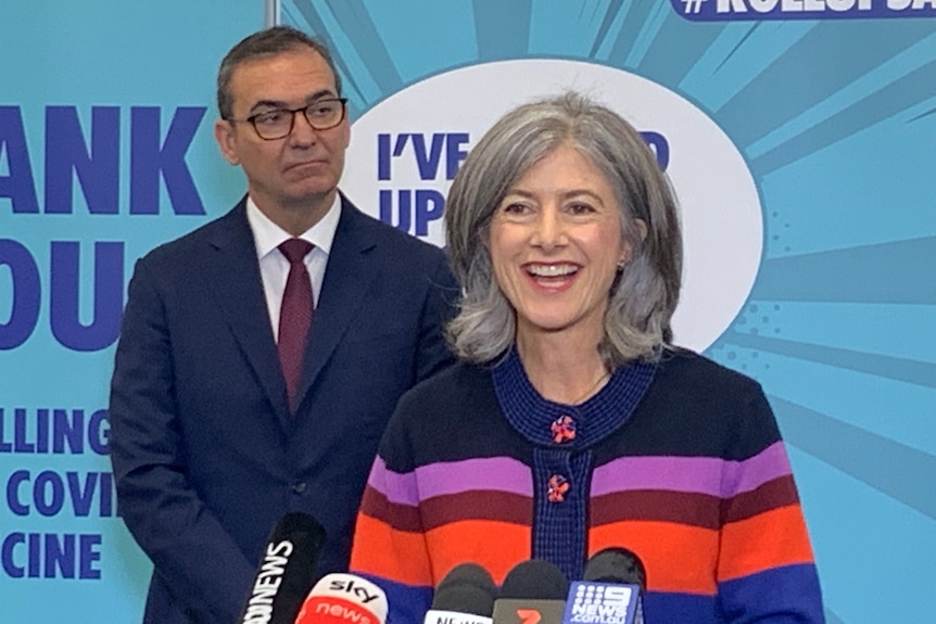 A smiling grey-haired woman wearing a colourful jumper in front of a man wearing a suit 